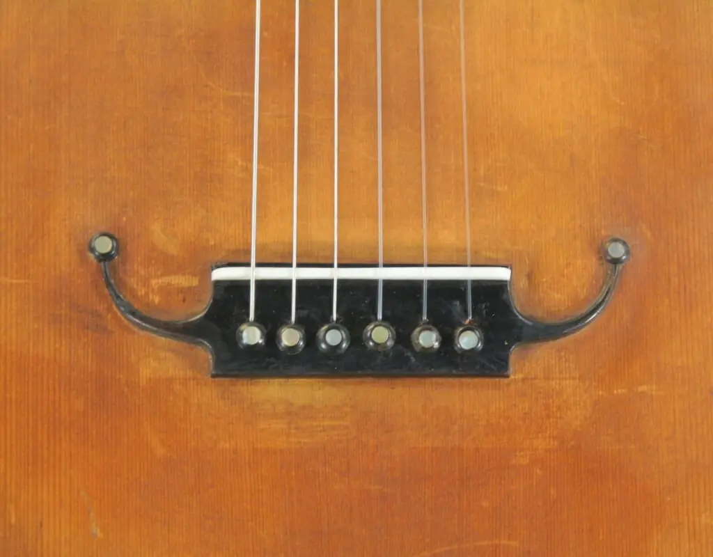 Nahaufnahme Des Stegs Der Französischen Romantikgitarre Von ~1860, Bei Dem Sechs Saiten An Einem Dunklen, Verzierten Steg Auf Einem Warmbraunen Holzkorpus Verankert Sind.