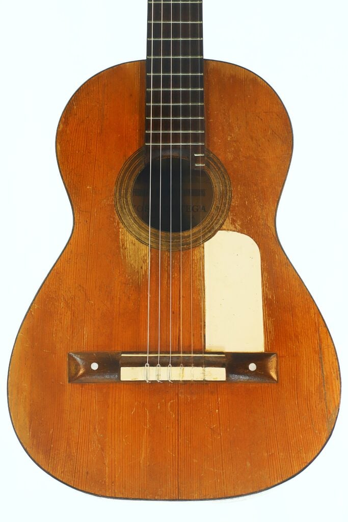 Eine Nahaufnahme der José Ortega ~1890, einer alten, abgenutzten Akustikgitarre mit sichtbaren Kratzern, sechs Saiten, einem runden Schallloch und einem hellen Schlagbrett auf der rechten Seite ihres Holzkörpers. Eine Nahaufnahme der José Ortega ~1890, einer alten, abgenutzten Akustikgitarre mit sichtbaren Kratzern, sechs Saiten, einem runden Schallloch und einem hellen Schlagbrett auf der rechten Seite ihres Holzkörpers.