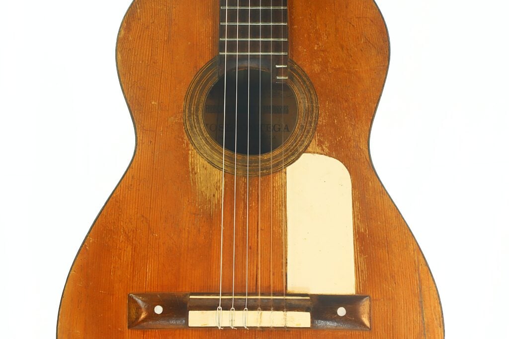 Nahaufnahme einer Akustikgitarre von José Ortega aus der Zeit um 1890, die die sichtbare Holzmaserung, die sechs Saiten, ein rundes Schallloch und einen rechteckigen, cremefarbenen Schlagbrettschutz unten rechts am Korpus zeigt. Nahaufnahme einer Akustikgitarre von José Ortega aus der Zeit um 1890, die die sichtbare Holzmaserung, die sechs Saiten, ein rundes Schallloch und einen rechteckigen, cremefarbenen Schlagbrettschutz unten rechts am Korpus zeigt.