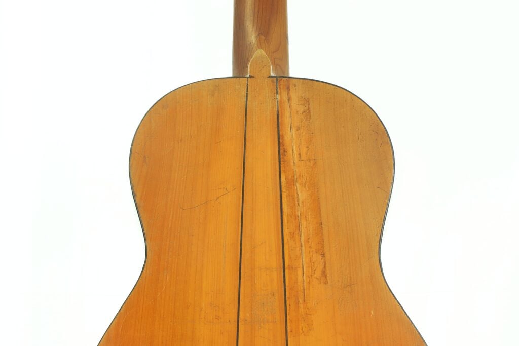 Nahaufnahme der Rückseite einer akustischen Gitarre von José Ortega (ca. 1890), die die Holzmaserung, die Mittelnaht und die Kratzer auf der Oberfläche vor einem schlichten weißen Hintergrund hervorhebt. Nahaufnahme der Rückseite einer akustischen Gitarre von José Ortega (ca. 1890), die die Holzmaserung, die Mittelnaht und die Kratzer auf der Oberfläche vor einem schlichten weißen Hintergrund hervorhebt.