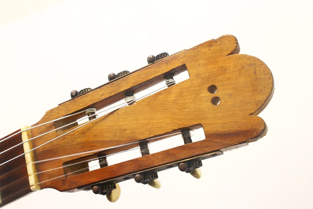 Nahaufnahme des Kopfstücks einer klassischen Gitarre von José Ortega aus der Zeit um 1890, mit sechs Stimmwirbeln, Nylonsaiten und einer Holzoberfläche mit zwei kleinen Löchern auf einer Seite. Nahaufnahme des Kopfstücks einer klassischen Gitarre von José Ortega aus der Zeit um 1890, mit sechs Stimmwirbeln, Nylonsaiten und einer Holzoberfläche mit zwei kleinen Löchern auf einer Seite.