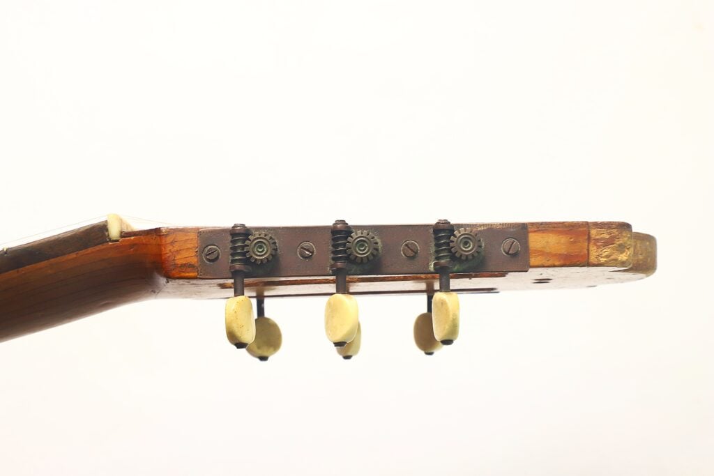 Nahaufnahme der Seitenansicht des Kopfes einer Holzgitarre von José Ortega (um 1890), die drei Stimmwirbel und Metallstimmmechaniken vor einem schlichten weißen Hintergrund zeigt. Nahaufnahme der Seitenansicht des Kopfes einer Holzgitarre von José Ortega (um 1890), die drei Stimmwirbel und Metallstimmmechaniken vor einem schlichten weißen Hintergrund zeigt.