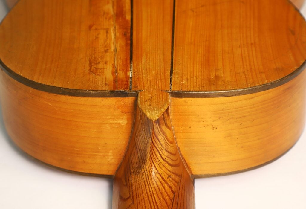 Nahaufnahme der Rückseite einer Akustikgitarre von José Ortega aus der Zeit um 1890, die den Holzkorpus, den Halsansatz und die sichtbaren Maserungen zeigt. Das Instrument weist Gebrauchsspuren und leichte Risse entlang der Fugen auf. Nahaufnahme der Rückseite einer Akustikgitarre von José Ortega aus der Zeit um 1890, die den Holzkorpus, den Halsansatz und die sichtbaren Maserungen zeigt. Das Instrument weist Gebrauchsspuren und leichte Risse entlang der Fugen auf.