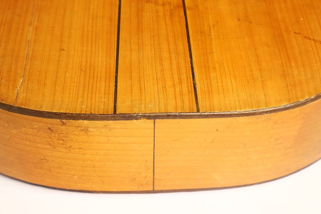 Nahaufnahme des Gitarrenkorpus von José Ortega (um 1890), auf der vertikale Risse und Kratzer auf der Holzoberfläche zu sehen sind, die Anzeichen von Abnutzung und Alterung deutlich machen. Nahaufnahme des Gitarrenkorpus von José Ortega (um 1890), auf der vertikale Risse und Kratzer auf der Holzoberfläche zu sehen sind, die Anzeichen von Abnutzung und Alterung deutlich machen.