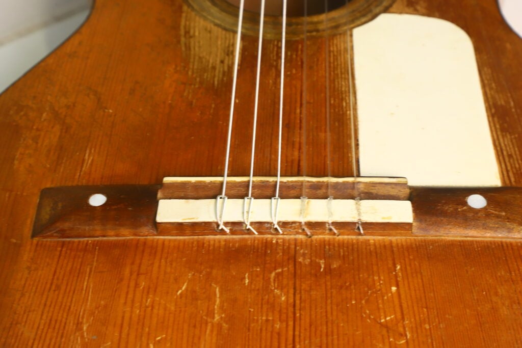 Nahaufnahme des Stegs und der Saiten der Akustikgitarre von José Ortega (ca. 1890), wobei die Holzmaserung, das Schlagbrett und die Gebrauchsspuren auf der Oberfläche des Instruments besonders hervorgehoben werden. Nahaufnahme des Stegs und der Saiten der Akustikgitarre von José Ortega (ca. 1890), wobei die Holzmaserung, das Schlagbrett und die Gebrauchsspuren auf der Oberfläche des Instruments besonders hervorgehoben werden.