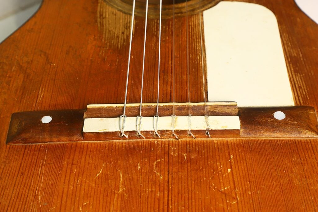 Nahaufnahme des Holzstegs und der Saiten der Akustikgitarre von José Ortega (ca. 1890), wobei die detaillierte, abgenutzte Holzoberfläche und ein weißes Schlagbrett auf der rechten Seite besonders hervorgehoben werden. Nahaufnahme des Holzstegs und der Saiten der Akustikgitarre von José Ortega (ca. 1890), wobei die detaillierte, abgenutzte Holzoberfläche und ein weißes Schlagbrett auf der rechten Seite besonders hervorgehoben werden.