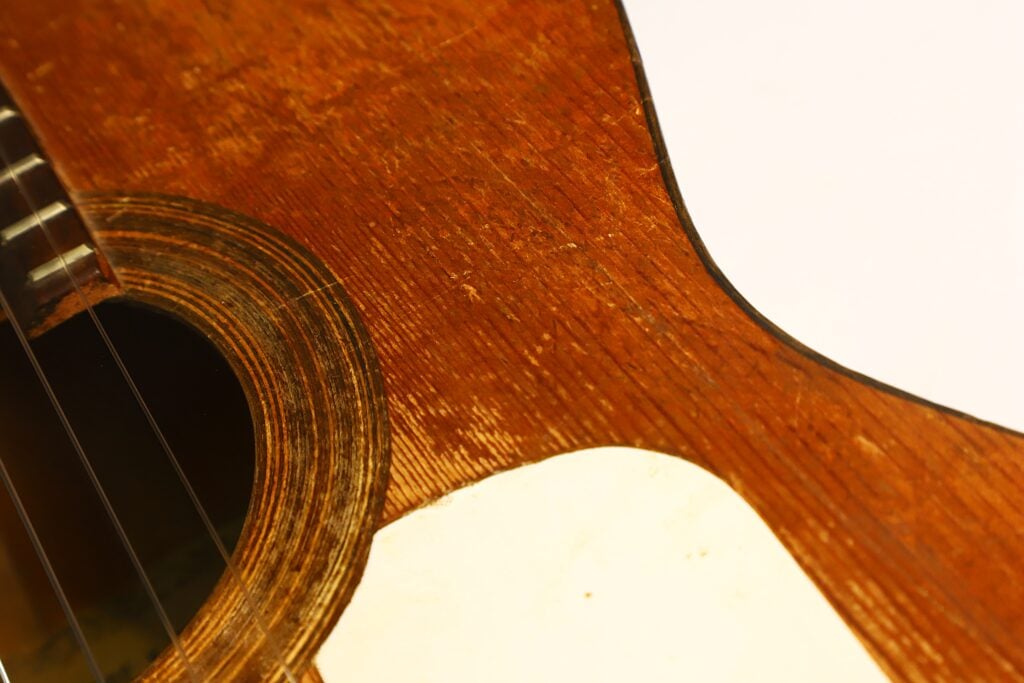Nahaufnahme des Korpus einer Akustikgitarre von José Ortega (ca. 1890), wobei das Schallloch, die Saiten, die Holzmaserung und ein Teil des weißen Schlagbretts vor einem schlichten Hintergrund hervorgehoben werden. Nahaufnahme des Korpus einer Akustikgitarre von José Ortega (ca. 1890), wobei das Schallloch, die Saiten, die Holzmaserung und ein Teil des weißen Schlagbretts vor einem schlichten Hintergrund hervorgehoben werden.