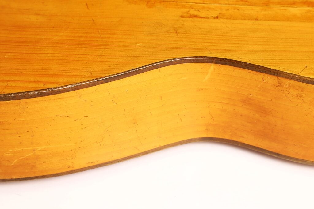 Nahaufnahme der Seite einer Akustikgitarre von José Ortega aus der Zeit um 1890, die die Holzmaserung, die geschwungene Form sowie sichtbare Kratzer und Abnutzungsspuren auf der Oberfläche hervorhebt. Nahaufnahme der Seite einer Akustikgitarre von José Ortega aus der Zeit um 1890, die die Holzmaserung, die geschwungene Form sowie sichtbare Kratzer und Abnutzungsspuren auf der Oberfläche hervorhebt.