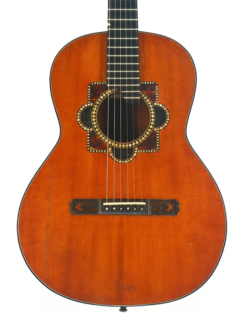 Eine Akustikgitarre von A. Klein, um 1920, mit einem rotbraunen Holzkorpus, aufwendig gestalteten Rosetten- und Schlagbrettmustern rund um das Schallloch, sechs Saiten und dekorativen Verzierungen am Steg, fotografiert vor weißem Hintergrund. Eine Akustikgitarre von A. Klein, um 1920, mit einem rotbraunen Holzkorpus, aufwendig gestalteten Rosetten- und Schlagbrettmustern rund um das Schallloch, sechs Saiten und dekorativen Verzierungen am Steg, fotografiert vor weißem Hintergrund.