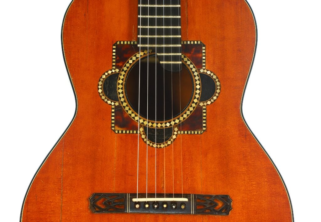 Nahaufnahme des Korpus der Akustikgitarre von A. Klein aus der Zeit um 1920, die die detailreichen geometrischen Einlegearbeiten rund um das Schallloch und den geschnitzten Holzsteg hervorhebt. Dieses Vintage-Instrument besticht durch eine satte, warmbraune Lackierung. Nahaufnahme des Korpus der Akustikgitarre von A. Klein aus der Zeit um 1920, die die detailreichen geometrischen Einlegearbeiten rund um das Schallloch und den geschnitzten Holzsteg hervorhebt. Dieses Vintage-Instrument besticht durch eine satte, warmbraune Lackierung.