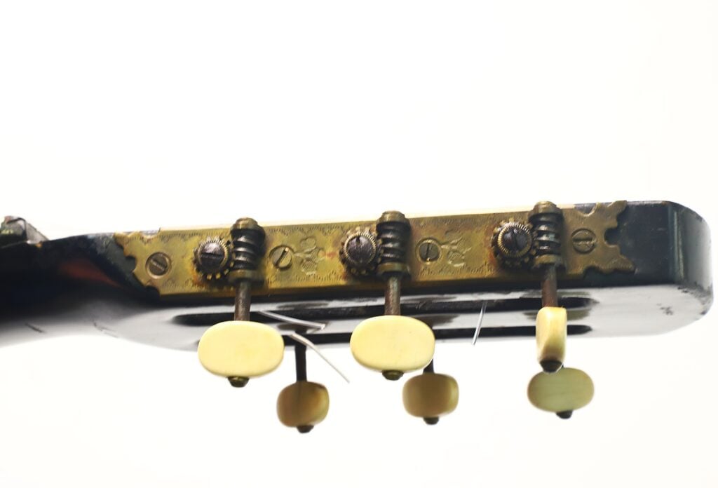 Nahaufnahme der Rückseite des Kopfes einer A. Klein-Gitarre aus der Zeit um 1920, die vier Stimmwirbel mit Metallzahnrädern und cremefarbenen Stimmknöpfen vor weißem Hintergrund zeigt. Nahaufnahme der Rückseite des Kopfes einer A. Klein-Gitarre aus der Zeit um 1920, die vier Stimmwirbel mit Metallzahnrädern und cremefarbenen Stimmknöpfen vor weißem Hintergrund zeigt.