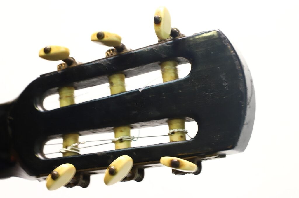 Nahaufnahme der Stimmwirbel und der Kopfplatte einer klassischen Gitarre von A. Klein aus der Zeit um 1920, auf der sechs weiße Stimmwirbel und Saiten vor weißem Hintergrund zu sehen sind. Nahaufnahme der Stimmwirbel und der Kopfplatte einer klassischen Gitarre von A. Klein aus der Zeit um 1920, auf der sechs weiße Stimmwirbel und Saiten vor weißem Hintergrund zu sehen sind.