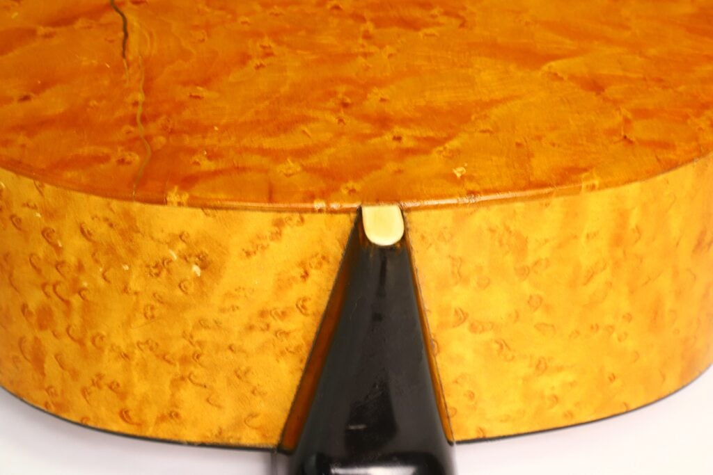 Nahaufnahme der Rückseite und des Randes der A. Klein-Akustikgitarre aus der Zeit um 1920, die die helle Holzmaserung und einen schwarzen Saitenhalter mit einem kleinen cremefarbenen Akzent hervorhebt. Nahaufnahme der Rückseite und des Randes der A. Klein-Akustikgitarre aus der Zeit um 1920, die die helle Holzmaserung und einen schwarzen Saitenhalter mit einem kleinen cremefarbenen Akzent hervorhebt.