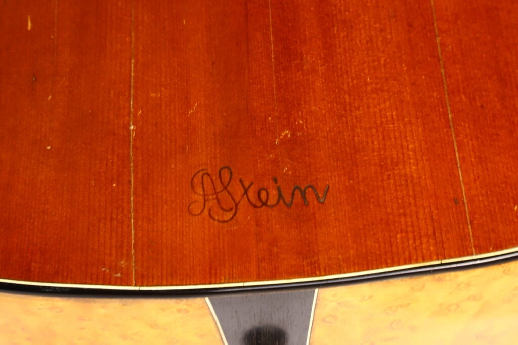 Nahaufnahme eines Gitarrenkorpus aus Holz, auf dessen Oberfläche nahe dem Rand und oberhalb der Einfassung die in schwarzer Tinte geschriebene, kursive Signatur „A. Klein“ zu sehen ist; es handelt sich um ein Modell aus der Zeit um 1920. Nahaufnahme eines Gitarrenkorpus aus Holz, auf dessen Oberfläche nahe dem Rand und oberhalb der Einfassung die in schwarzer Tinte geschriebene, kursive Signatur „A. Klein“ zu sehen ist; es handelt sich um ein Modell aus der Zeit um 1920.
