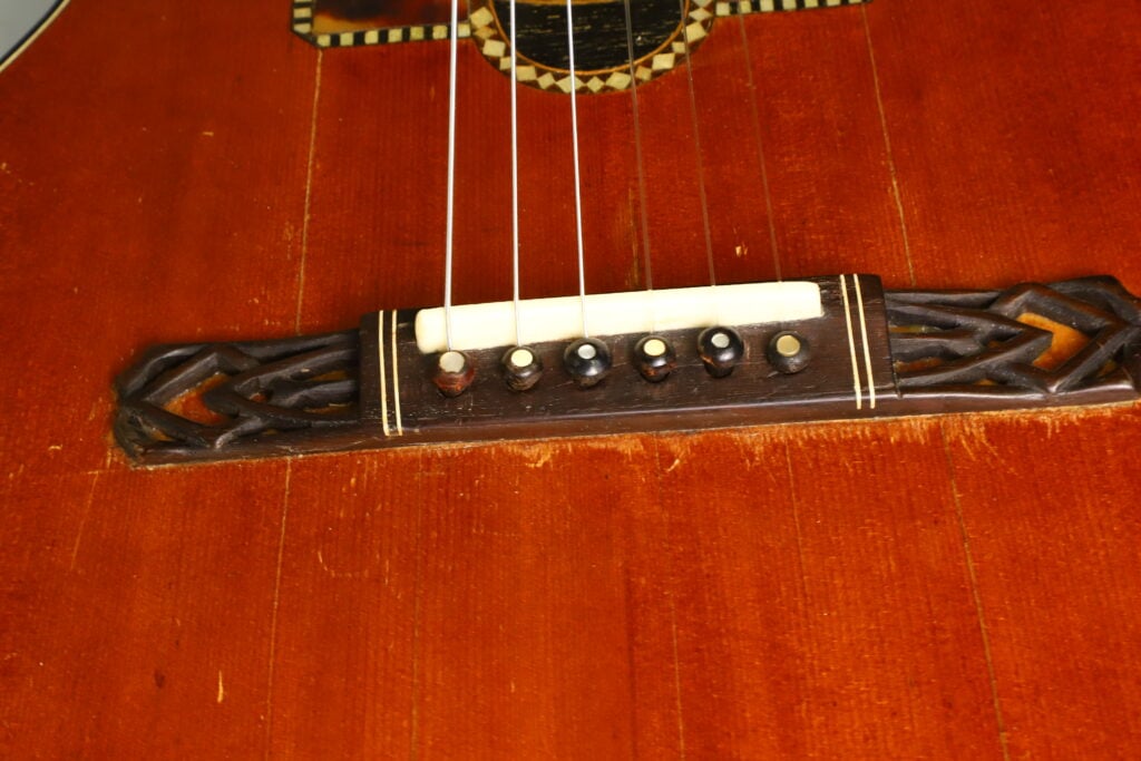 Nahaufnahme des Stegs einer A. Klein-Akustikgitarre aus der Zeit um 1920, auf der sechs Stegstifte, Saiten und aufwendig geschnitzte Holzdetails auf dem rotbraunen Korpus zu sehen sind. Nahaufnahme des Stegs einer A. Klein-Akustikgitarre aus der Zeit um 1920, auf der sechs Stegstifte, Saiten und aufwendig geschnitzte Holzdetails auf dem rotbraunen Korpus zu sehen sind.