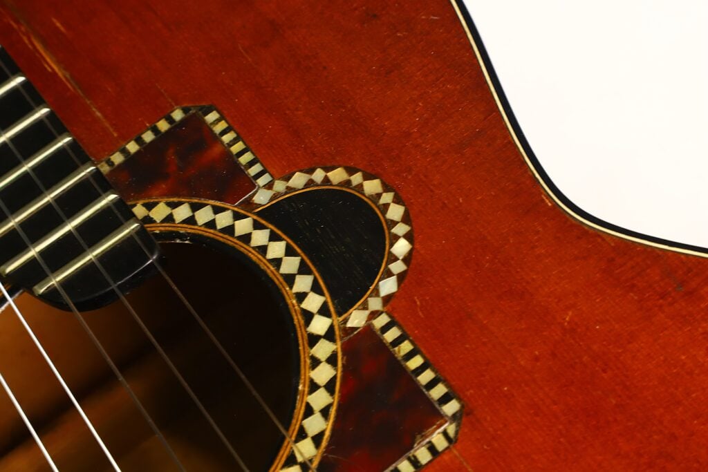 Nahaufnahme des Schalllochs der Akustikgitarre von A. Klein (um 1920) mit einem aufwendigen geometrischen Einlegemuster in Schwarz und Weiß, dunklen Saiten und einem satten rotbraunen Holzkorpus. Nahaufnahme des Schalllochs der Akustikgitarre von A. Klein (um 1920) mit einem aufwendigen geometrischen Einlegemuster in Schwarz und Weiß, dunklen Saiten und einem satten rotbraunen Holzkorpus.