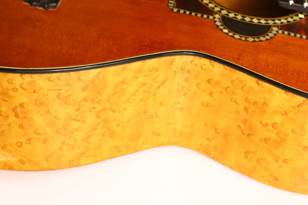 Nahaufnahme der Seite einer A. Klein-Akustikgitarre aus der Zeit um 1920, die detaillierte Holzmaserungen und eine glänzende Lackierung zeigt, wobei ein Teil des Korpus und die Zierleisten zu sehen sind. Nahaufnahme der Seite einer A. Klein-Akustikgitarre aus der Zeit um 1920, die detaillierte Holzmaserungen und eine glänzende Lackierung zeigt, wobei ein Teil des Korpus und die Zierleisten zu sehen sind.