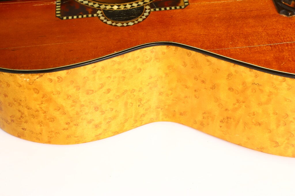 Nahaufnahme der Seite einer A. Klein-Gitarre aus der Zeit um 1920, die die hellgelbe, maserige Holzmaserung hervorhebt; darüber ist vor weißem Hintergrund ein Teil der dunkleren, verzierten Resonanzdecke zu sehen. Nahaufnahme der Seite einer A. Klein-Gitarre aus der Zeit um 1920, die die hellgelbe, maserige Holzmaserung hervorhebt; darüber ist vor weißem Hintergrund ein Teil der dunkleren, verzierten Resonanzdecke zu sehen.