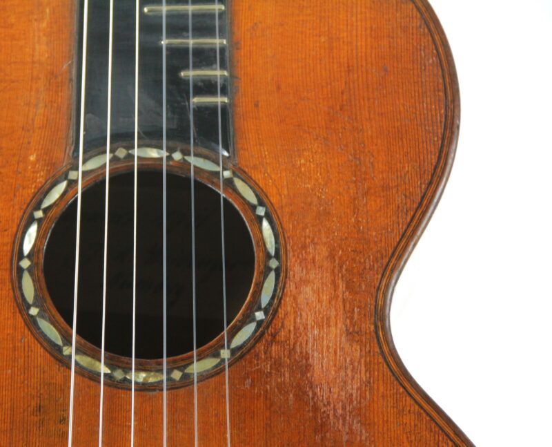 Close-up of the Stauffer Style ~1830 Romantic Guitar, showing the sound hole with a decorative rosette, several strings, and the upper right edge of the wooden body against a white background.