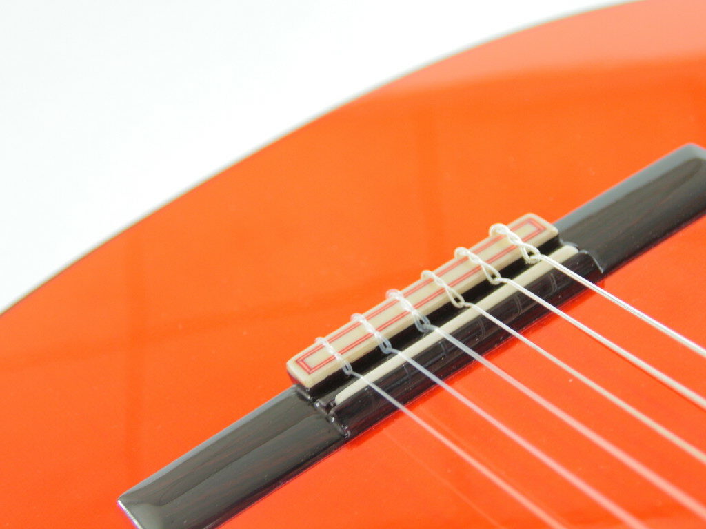 Nahaufnahme Der Saiten Und Des Stegs Einer Hermanos Conde 1999 Paco De Lucia Spezial Flamenco Gitarre. Hervorzuheben Sind Die Feinen Saitendetails Und Der Glänzende Orangefarbene Korpus Vor Weißem Hintergrund.