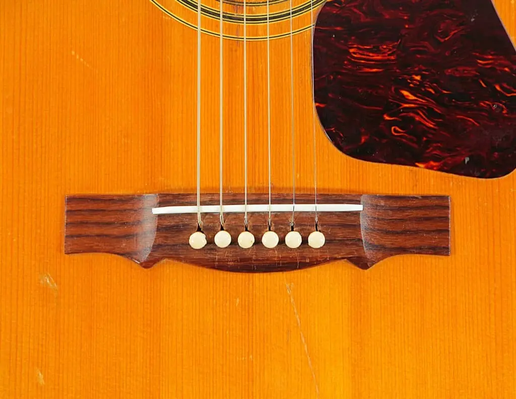 Close-Up Of The Guild D-44 1967 Acoustic Guitar Bridge And Strings On Its Wooden Body, With A Dark Brown Pickguard Visible In The Upper Right.