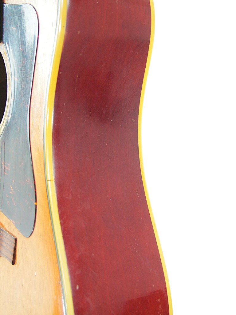 A Close-Up Side View Of The Guild D-44 1967 Acoustic Guitar Displays Its Curved, Reddish-Brown Wooden Body With A Light Yellow Edge, Plus Part Of The Sound Hole And Pickguard On The Left Against A White Background.