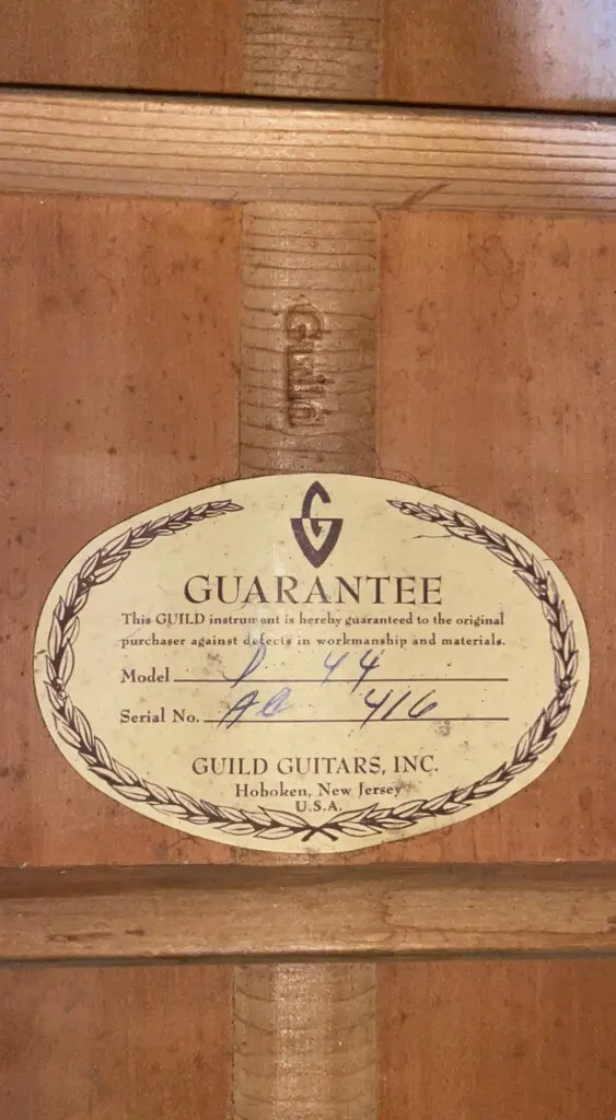 An Oval Label Inside The Wooden Body Of The Guild D-44 1967 Acoustic Guitar Reads Guarantee And Displays Guild Guitars, Inc. Info, With Handwritten Model And Serial Numbers. The Guild Logo Is Printed Above The Text.