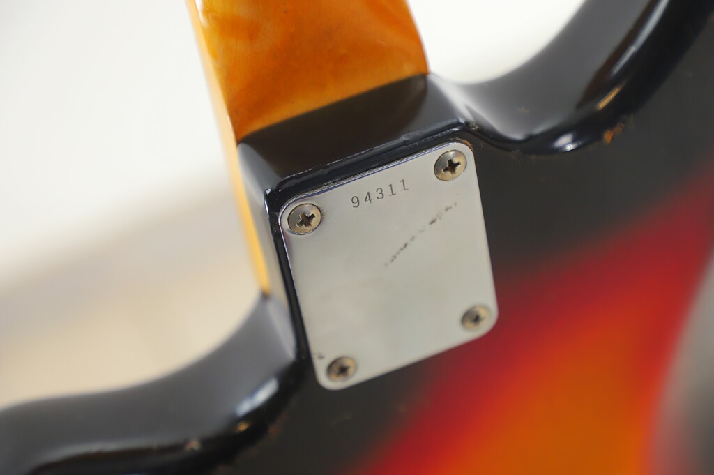 Close-Up Of A Fender Jazzmaster 1963 Electric Guitar Neck Joint, Showing A Four-Screw Metal Plate Engraved With 94311. The Classic Sunburst Finish Highlights The Vintage Design.