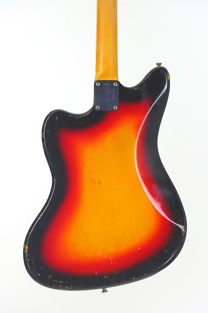 A Sunburst Fender Jazzmaster 1963 Electric Guitar Is Shown From The Back, Featuring A Worn Vintage Finish With Visible Scratches And Scuffs, Plus A Natural Wood Neck, All Set Against A Plain White Background.