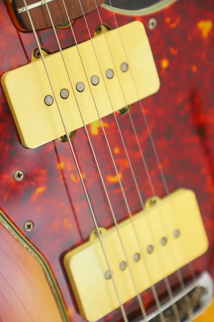 Close-Up Of A Fender Jazzmaster 1963 Electric Guitar’s Body Showing Two Yellow Single Coil Pickups, Metal Strings, And A Red Tortoiseshell Pickguard. The Wood Grain And Finish Wear Are Visible.