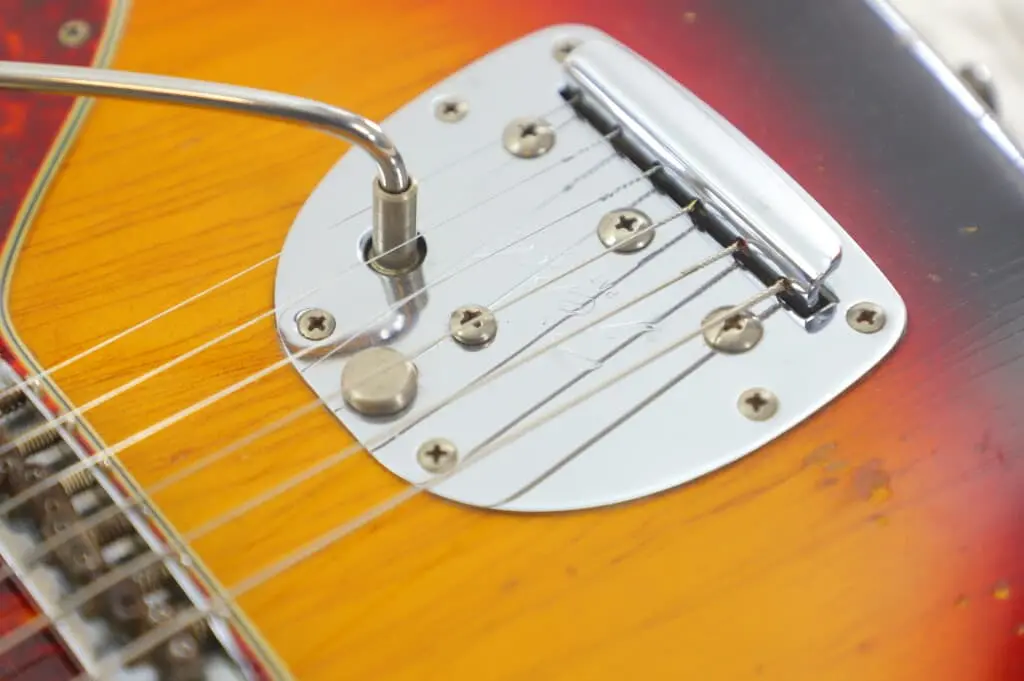 Close-Up Of The Fender Jazzmaster 1963 Electric Guitar’s Bridge And Tremolo Arm, Showcasing Its Metal Parts, Strings, And The Vivid Orange-Red Sunburst Wood Finish.