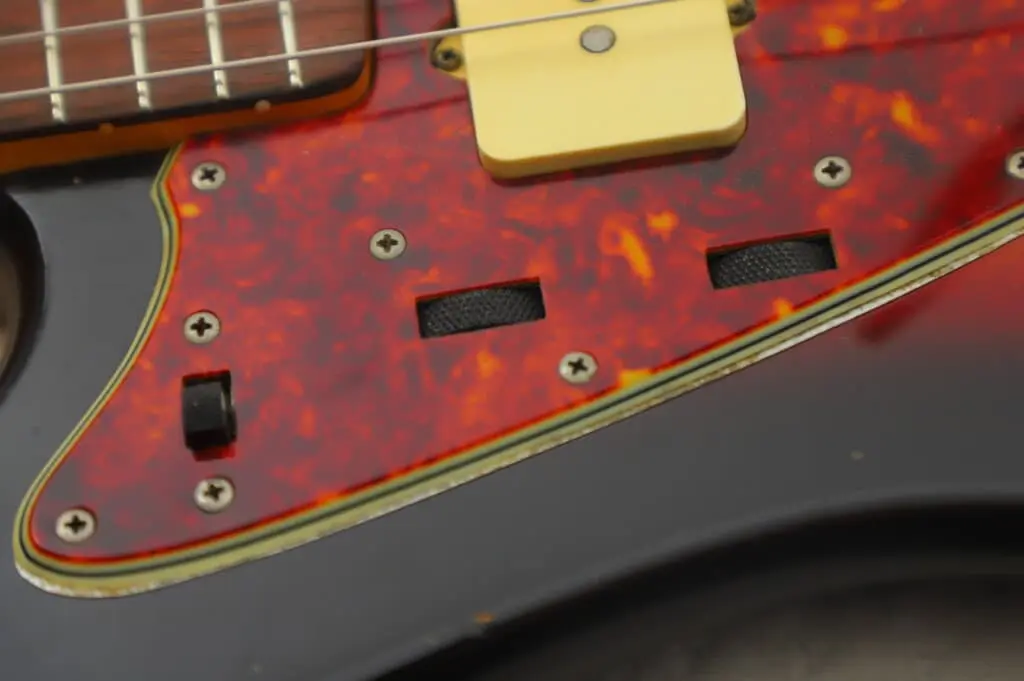 Close-Up Of A Fender Jazzmaster 1963 Electric Guitar’s Body Featuring A Red Tortoiseshell Pickguard, Two Empty Rectangular Switch Slots, A Small Black Switch, And Part Of The Strings And Pickups At The Top.