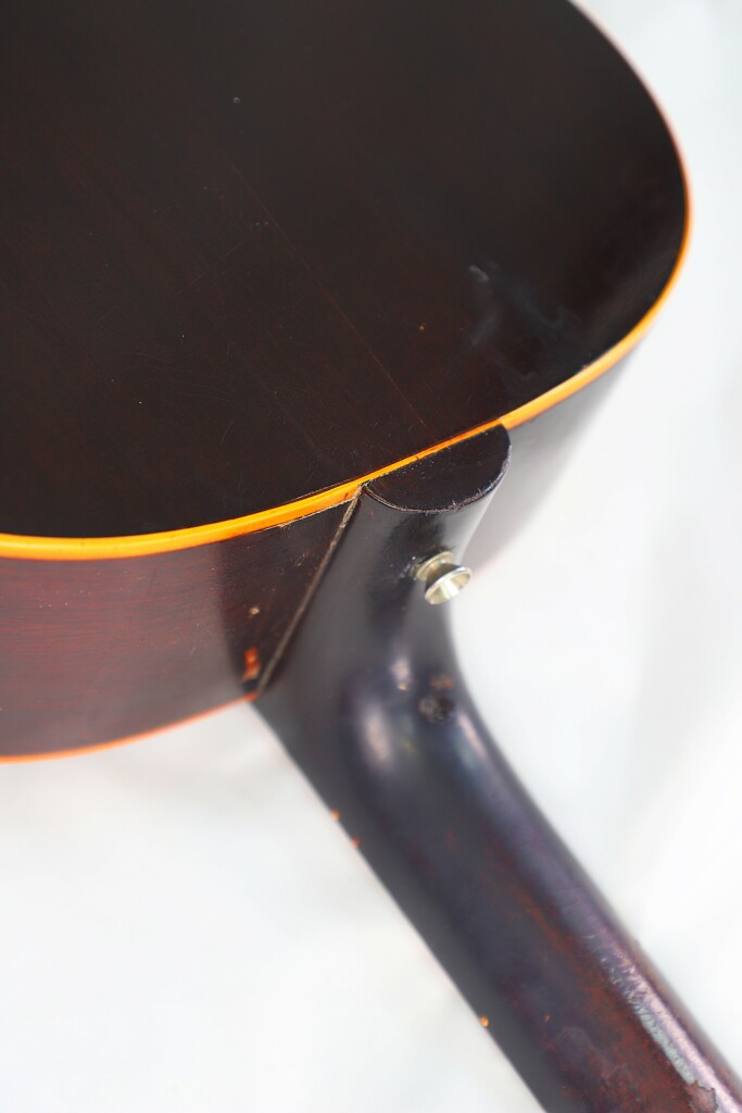Close-Up Of The Back And Neck Joint Of A Dark Brown Gibson J-45 1955 Acoustic Guitar, Featuring A Glossy Finish And Yellow Edging, Set Against A White Background.