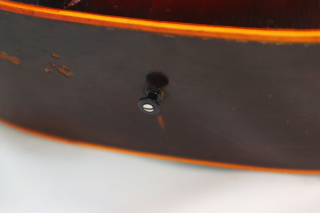 Close-Up Of The Side Of A Worn Gibson J-45 1955 Acoustic Guitar, Featuring A Black Strap Button On The Wood Near The Edge, With Visible Scratches And Chipped Areas.