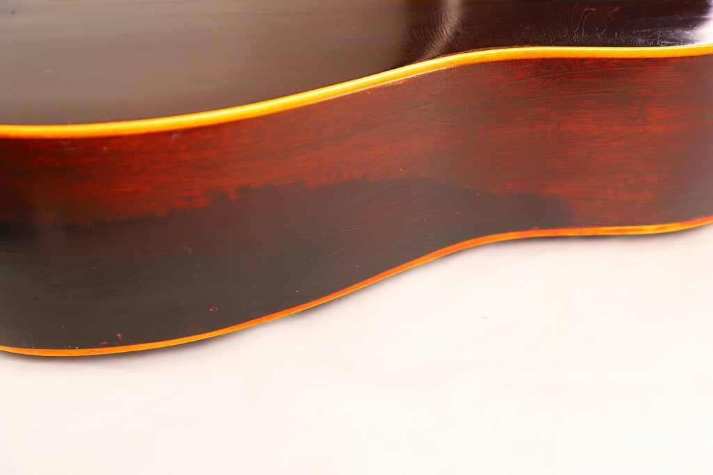 Close-Up Of The Gibson J-45 1955 Acoustic Guitars Side, Showing Its Dark Wood Finish And Yellowish Trim, With Smooth Curves Highlighted Against A Light Background.