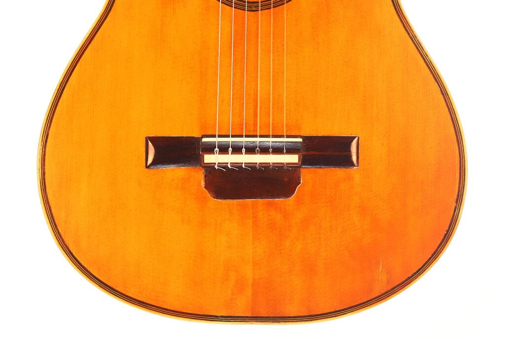 Nahaufnahme der unteren Hälfte einer klassischen Akustikgitarre von Francisco Manuel Fleta aus dem Jahr 1962. Hervorzuheben sind die sechs Saiten, die glänzende Holzoberfläche und die detaillierte Verarbeitung von Korpus und Steg. Nahaufnahme der unteren Hälfte einer klassischen Akustikgitarre von Francisco Manuel Fleta aus dem Jahr 1962. Hervorzuheben sind die sechs Saiten, die glänzende Holzoberfläche und die detaillierte Verarbeitung von Korpus und Steg.