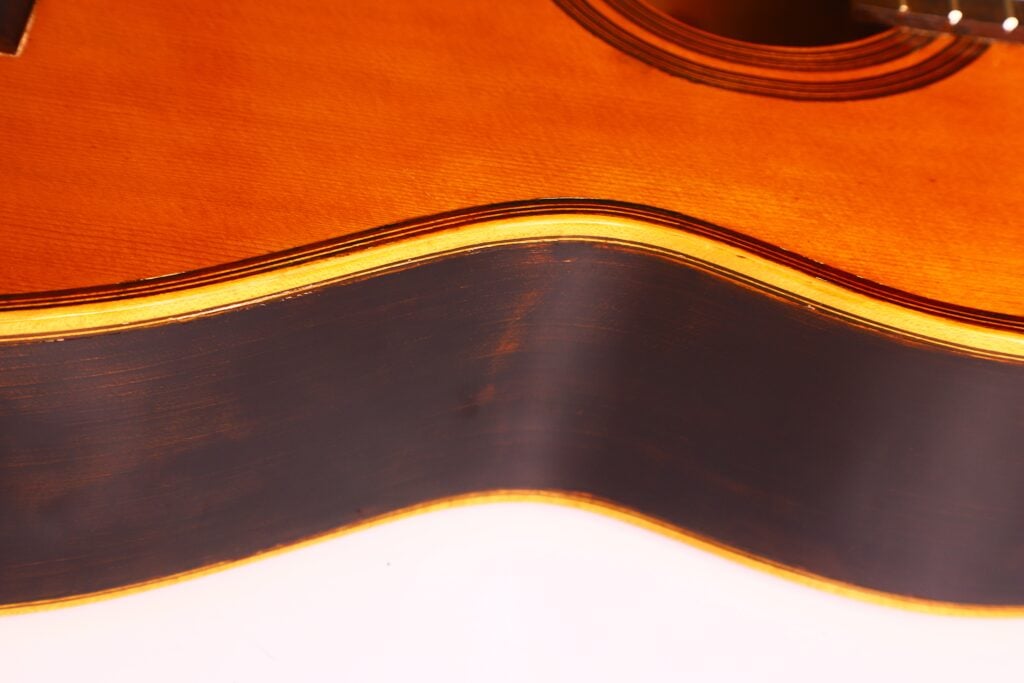 Nahaufnahme der Seite und der Decke einer 1962er Akustikgitarre von Francisco Manuel Fleta. Hervorzuheben sind die Holzmaserung, die Rundungen des Korpus und die glänzende warm-braune Lackierung. Nahaufnahme der Seite und der Decke einer 1962er Akustikgitarre von Francisco Manuel Fleta. Hervorzuheben sind die Holzmaserung, die Rundungen des Korpus und die glänzende warm-braune Lackierung.