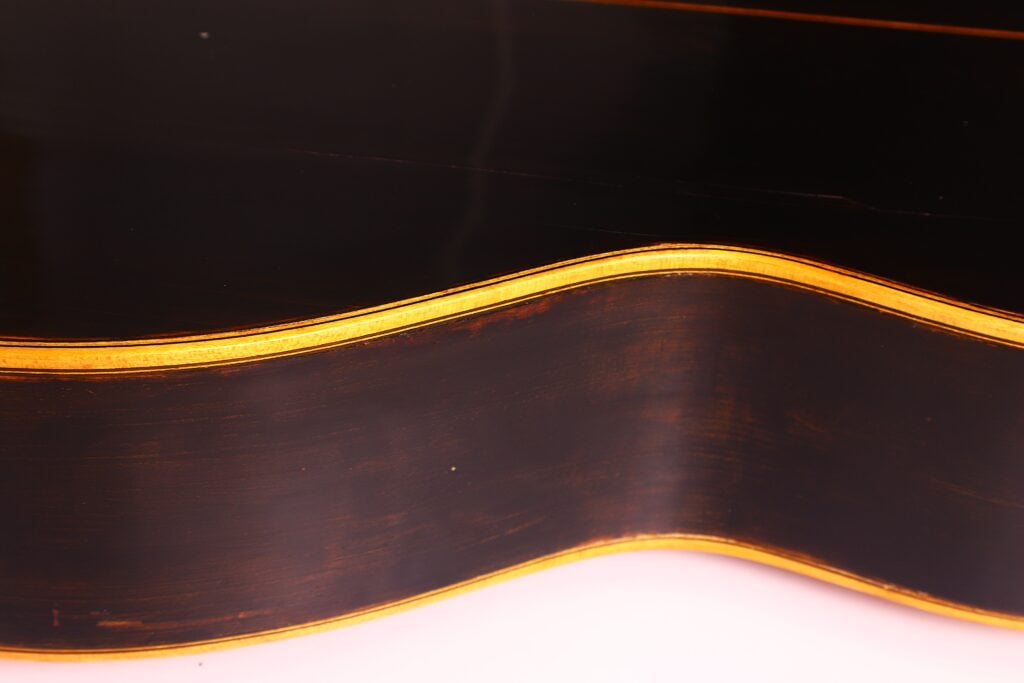 Nahaufnahme der gewölbten Seite einer Akustikgitarre von Francisco Manuel Fleta aus dem Jahr 1962 mit dunkler Hochglanzlackierung und goldenen Verzierungen, die die Holzmaserung und die Konturen vor einem hellen Hintergrund zur Geltung bringen. Nahaufnahme der gewölbten Seite einer Akustikgitarre von Francisco Manuel Fleta aus dem Jahr 1962 mit dunkler Hochglanzlackierung und goldenen Verzierungen, die die Holzmaserung und die Konturen vor einem hellen Hintergrund zur Geltung bringen.