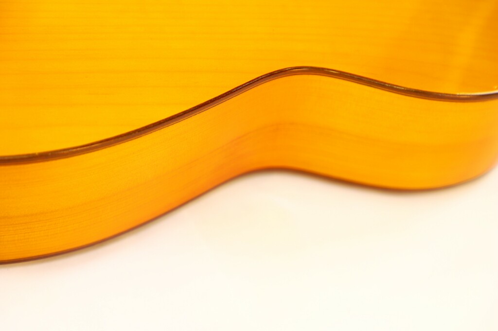 Close-Up Of The Side Of A Manuel G. Mayoral 1980 Flamenco Guitar, Featuring Its Smooth, Curved Wooden Body And Glossy Light Brown Finish Against A White Background.