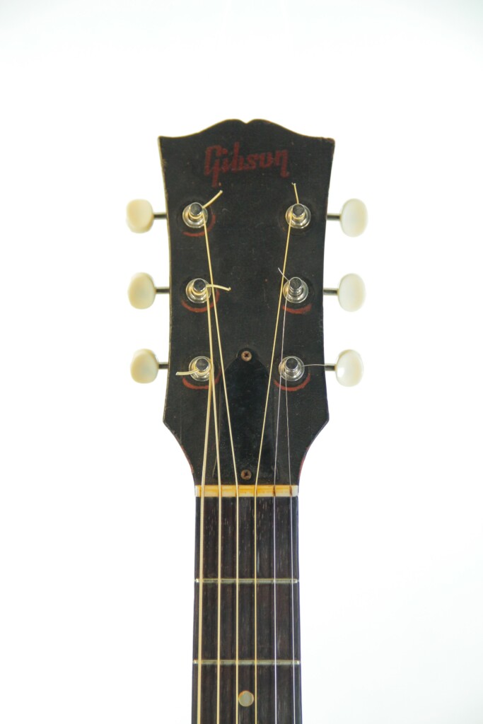 Close-Up Of The Gibson J-45 1955 Acoustic Guitar Headstock, Featuring Six Tuning Pegs And The Gibson Logo On Top, With Part Of The Fretboard Below Against A White Background.