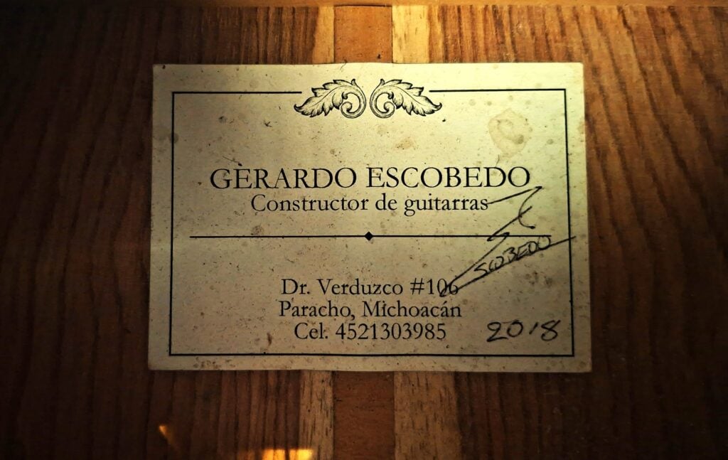 Ein rechteckiges Papieretikett im Inneren einer Flamenco-Gitarre von Gerardo Escobedo aus dem Jahr 2018, auf dem die Adresse des Herstellers, seine Telefonnummer, eine Unterschrift und das Jahr 2018 angegeben sind. Ein rechteckiges Papieretikett im Inneren einer Flamenco-Gitarre von Gerardo Escobedo aus dem Jahr 2018, auf dem die Adresse des Herstellers, seine Telefonnummer, eine Unterschrift und das Jahr 2018 angegeben sind.
