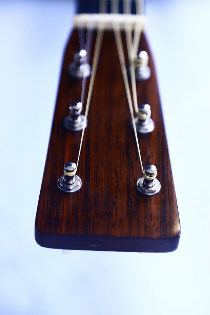 Close-Up Of The Martin Om-28 1931 Acoustic Guitar Headstock, Featuring Six Tuning Pegs, Metal Strings, And Visible Wood Grain Against A Soft Blue Blurred Background.