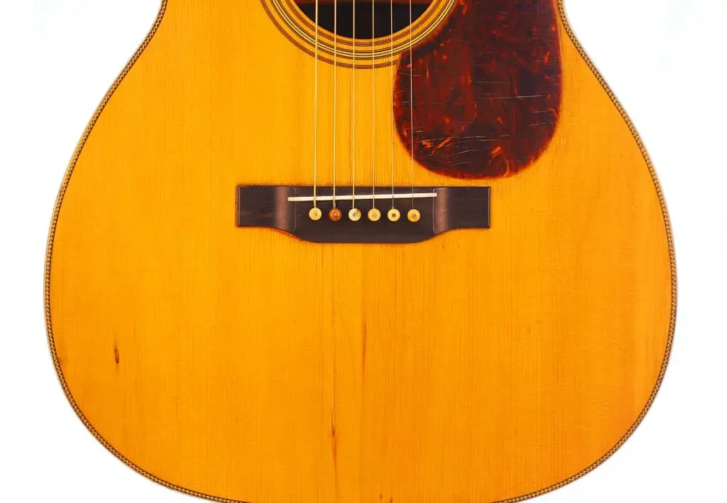 Close-Up Of The Martin Om-28 1931 Acoustic Guitar Body, Featuring Its Wooden Surface, Six Strings, Dark Bridge, And A Brown Tortoiseshell-Style Pickguard On The Right.