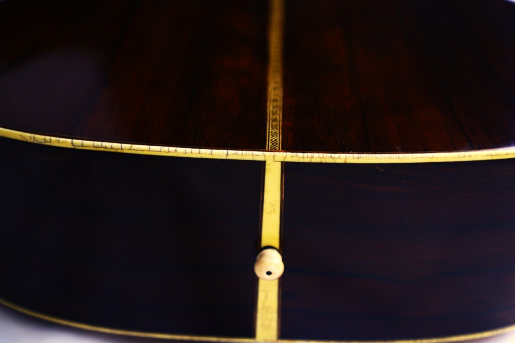Close-Up Of The Back And Side Of The Martin Om-28 1931 Acoustic Guitar, Showing Its Dark Wood Finish, Decorative Yellow Inlay, And A Small Wooden End Pin At The Bottom.