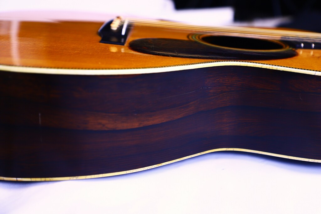 Close-Up Side View Of The Martin Om-28 1931 Acoustic Guitar On A White Surface, Showcasing Its Wood Grain And Glossy Finish, With Part Of The Strings And Sound Hole Visible.