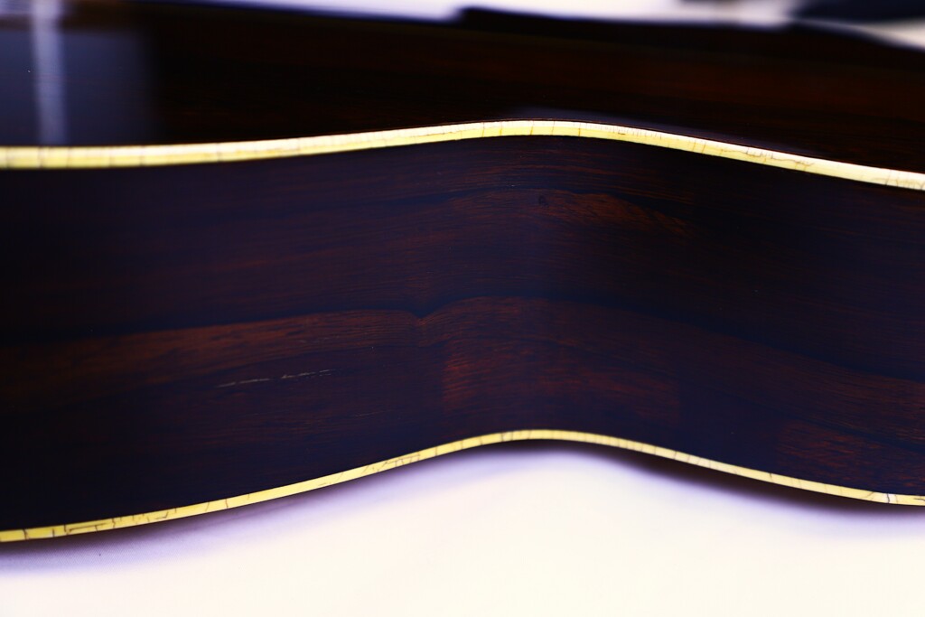 Close-Up Of The Martin Om-28 1931 Acoustic Guitar’s Side, Highlighting Its Smooth, Dark Wood Grain, Glossy Finish, And Cream-Colored Binding Along The Edges.