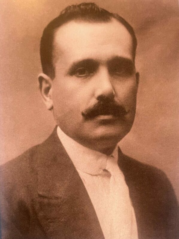 Ein Sepiafarbenes Porträt Eines Mannes Mit Dickem Schnurrbart, Kurzen Dunklen Haaren Und Einer Anzugsjacke Über Einem Weißen Hemd. Er Blickt Direkt In Die Kamera Und Hat Einen Neutralen Ausdruck.