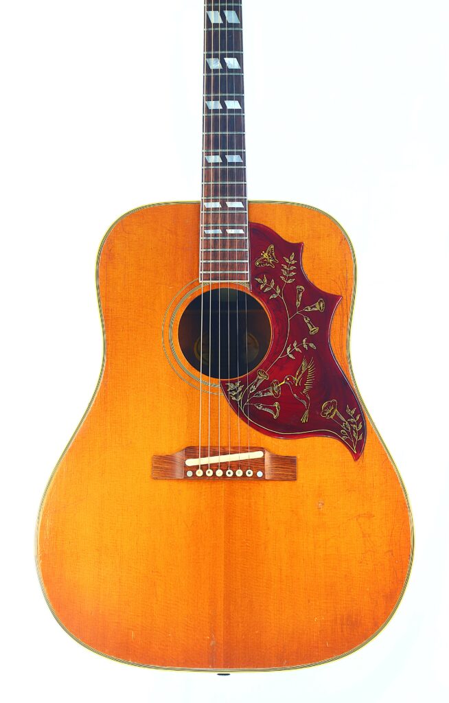 Die Gibson Hummingbird 1966 Akustik Gitarre Verfügt Über Ein Naturholz-Finish, Dekorative Griffbretteinlagen Und Ein Rotes Schlagbrett, Das Mit Blumen- Und Vogelmotiven Verziert Ist.