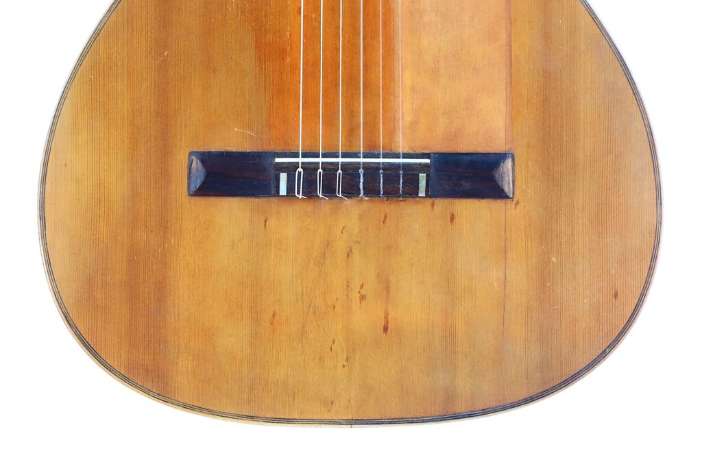Close-Up Of The Lower Half Of A Salvador Ibanez ~1900 Classical Guitar, Highlighting Its Wooden Body, Six Strings, And Dark Bridge Against A White Background.