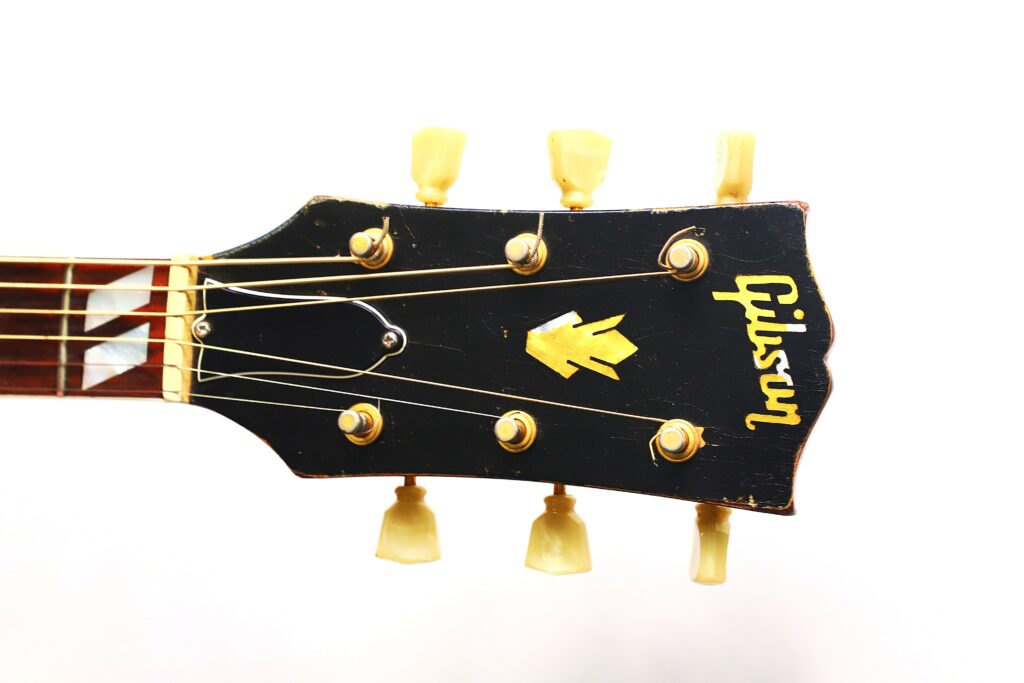 Nahaufnahme Der Kopfplatte Einer Gibson Hummingbird 1966 Akustik Gitarre Mit Schwarzem Finish, Gelben Stimmwirbeln Und Dem Gibson Logo In Gelb Auf Weißem Hintergrund.